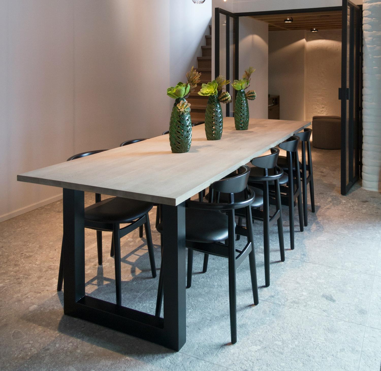 Lange massief houten tafel met zwart stalen onderstel, zwarte stoelen en groene vazen in modern interieur